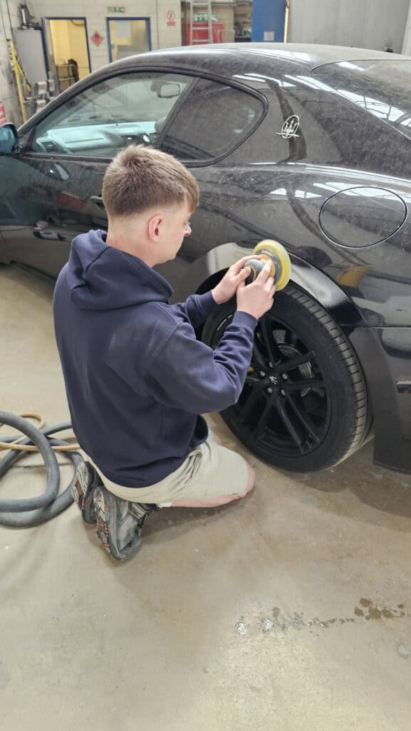Technician repairing a black sports car with a polishing tool in an auto repair shop.